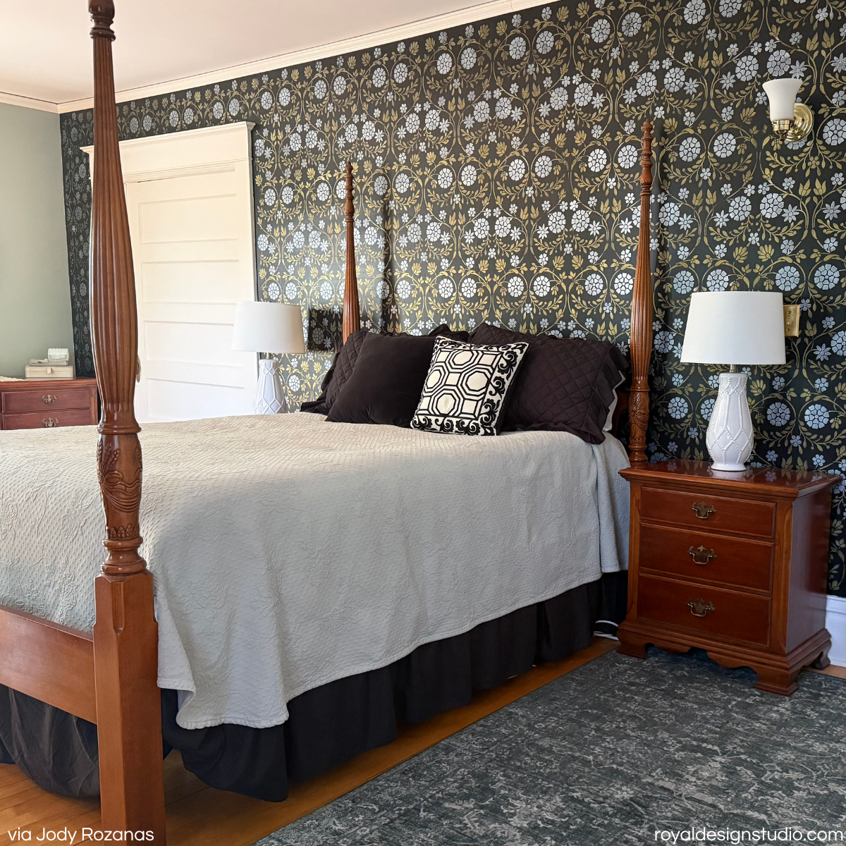 A styled bedroom featuring a wooden four-poster bed with a light beige textured quilt, dark brown pillows, and a black and white patterned accent pillow. The bed is set against a wall covered in dark green and white floral hand stenciled pattern