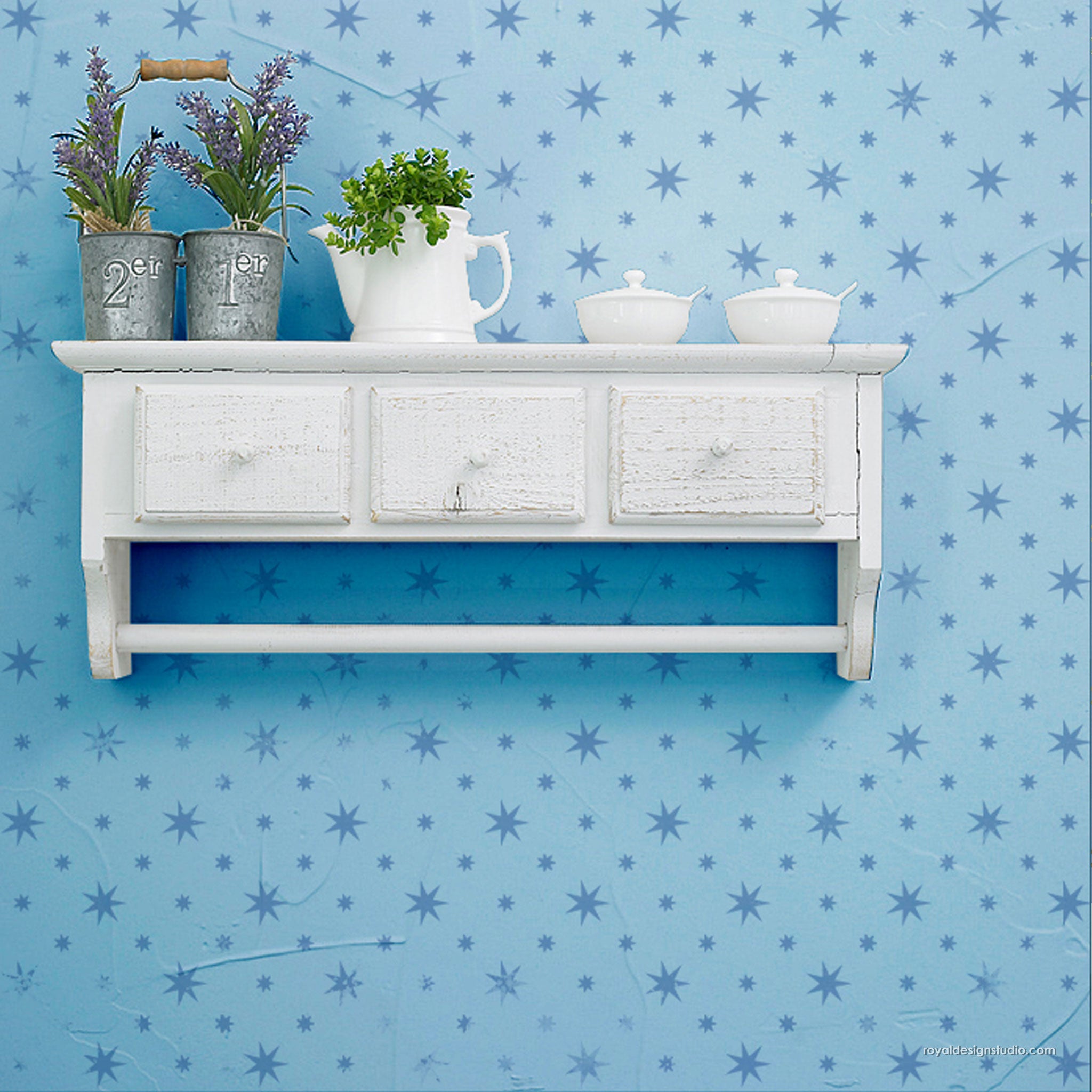 A white rustic wall-mounted shelf with three drawers and a lower bar, styled with lavender plants, a white pitcher, and three white sugar bowls against a blue star-patterned wall background.