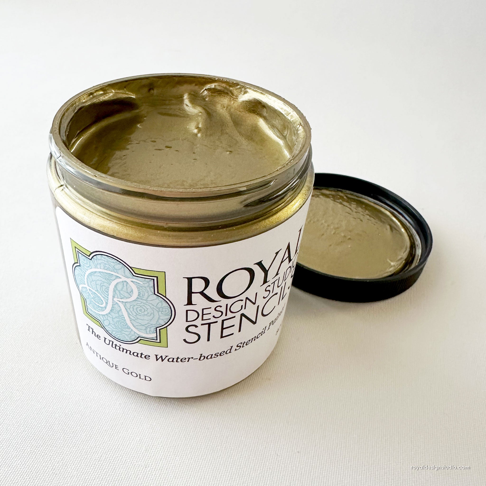 An open jar of Royal Design Studio Stencil Paint in 'Antique Gold' with its black lid resting beside it on a light off-white background.