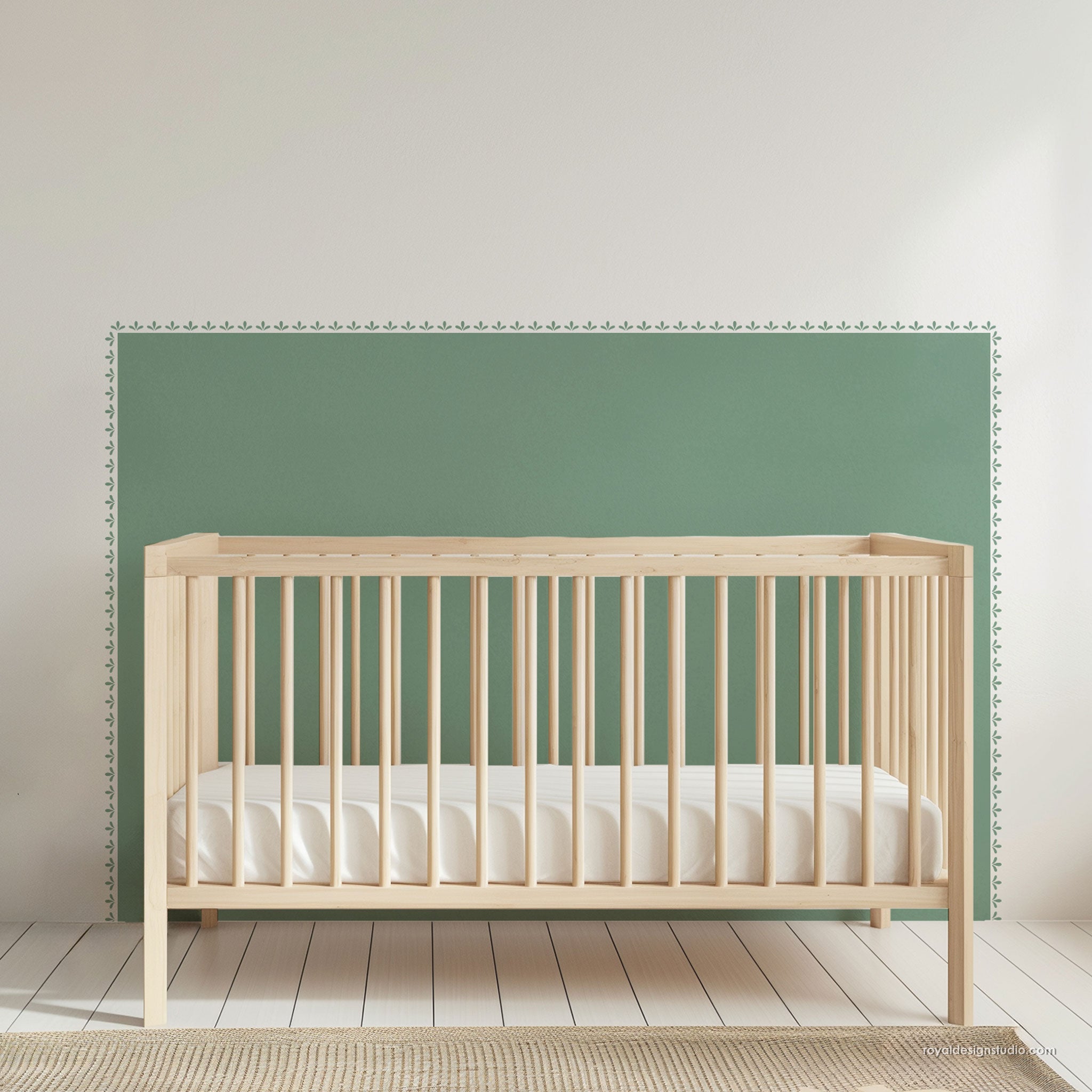 A light natural wood baby crib with white mattress sheets, positioned in front of a sage green painted wall with a white wooden floor.