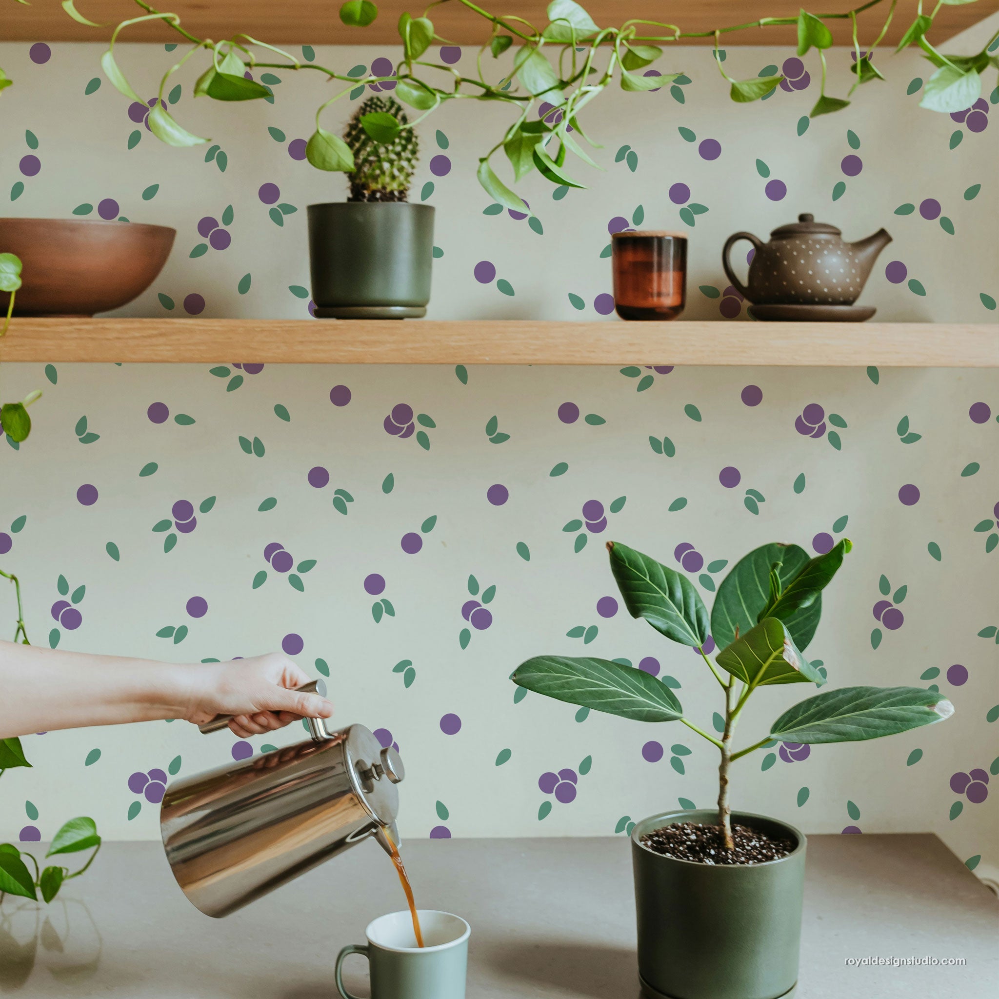 A lifestyle scene showing a person pouring coffee from a stainless steel pitcher into a mug, set against a wall with a repeating purple and green berry pattern on a cream background. Light wood shelves with plants and a teapot are visible above.