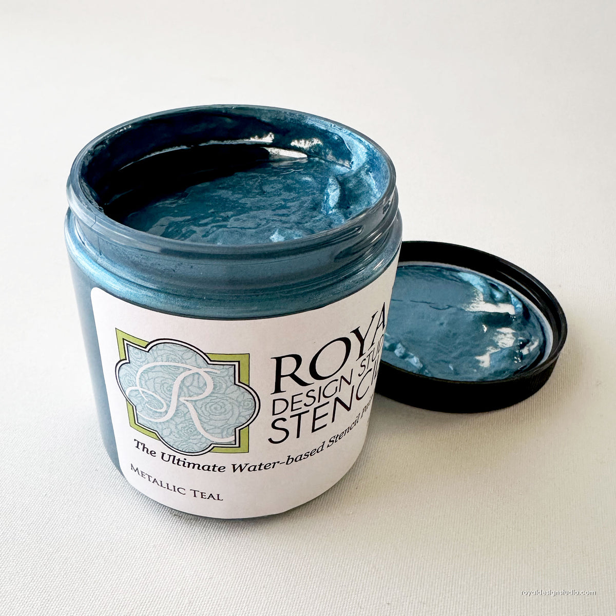 An open jar of Royal Design Studio metallic teal stenciling paint with its black lid resting beside it on a light beige textured background.