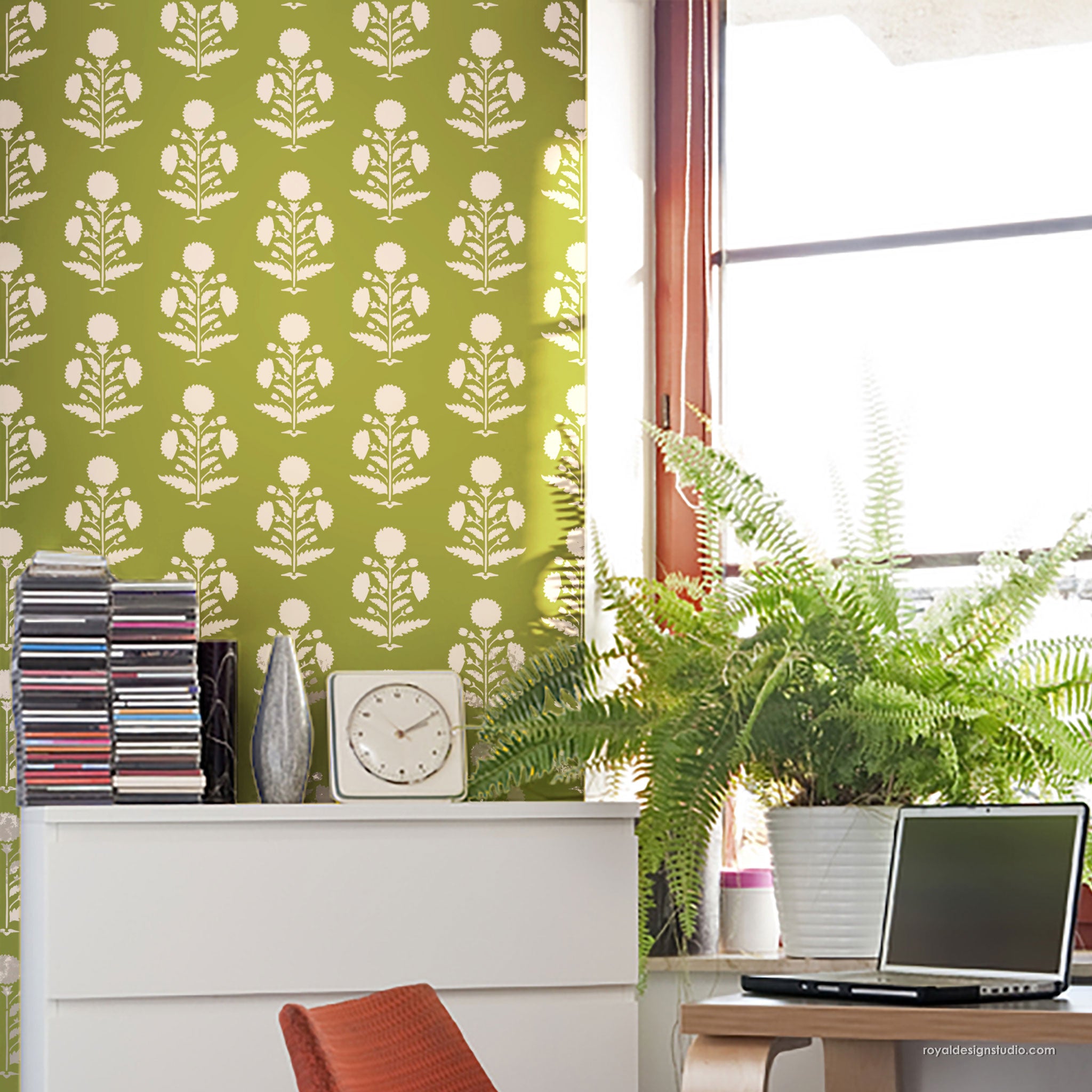 Green and white floral patterned wall in a styled room setting with a white cabinet, stack of CDs, a clock, a potted fern, and a laptop.