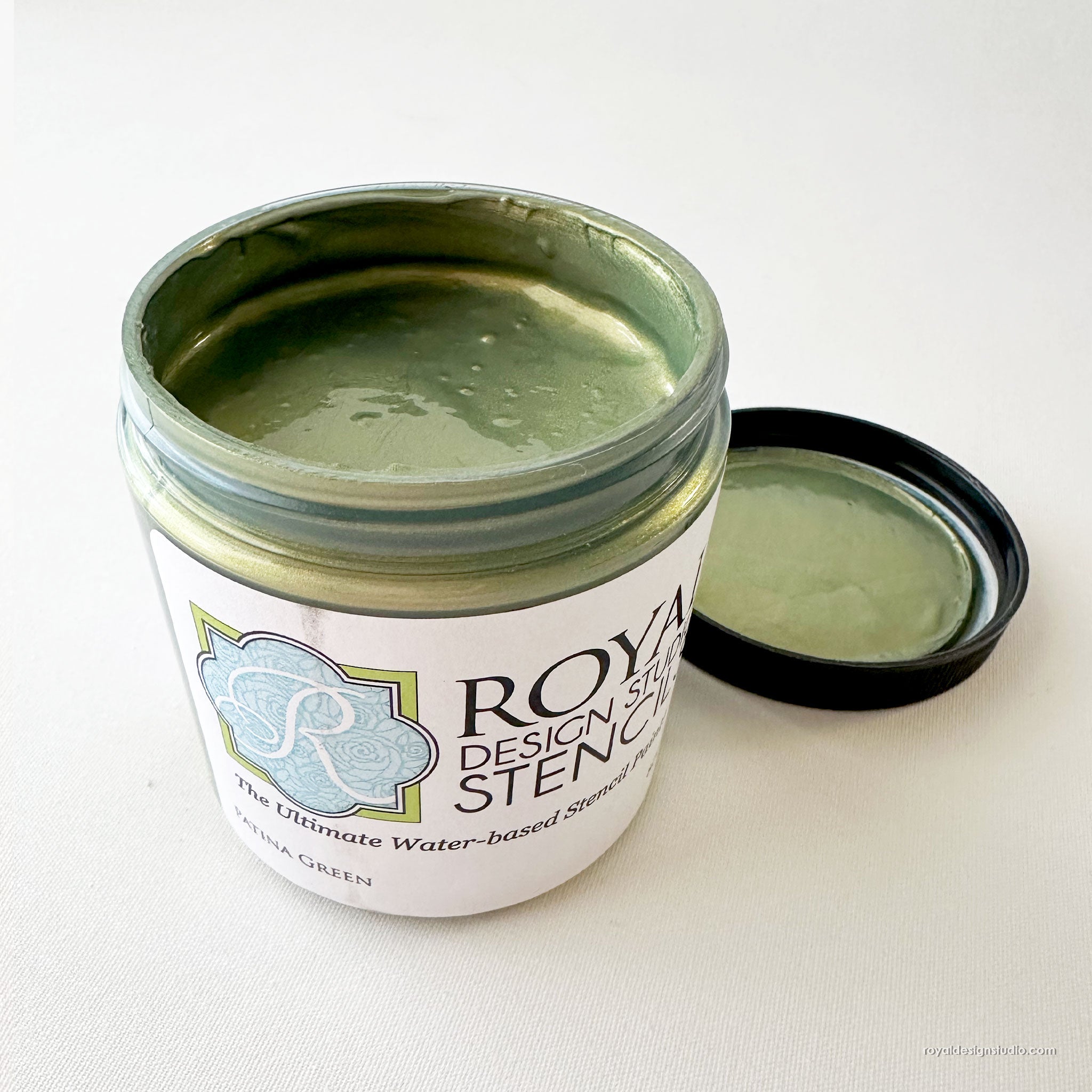 An open jar of Royal Design Studio Stencil in the color 'Patina Green' shows the textured green stencil paste inside, with its black lid resting beside it on a light off-white background.