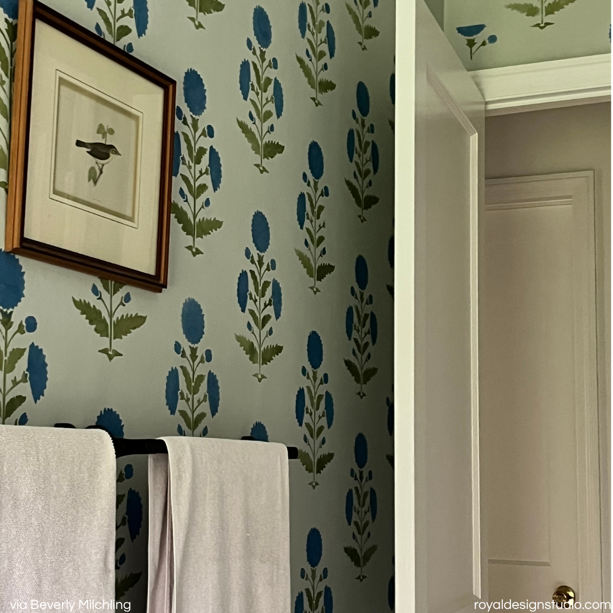Blue and green floral block-print style wall stencil in a bathroom setting, shown with a framed bird print on the wall, white towels on a black towel bar, and an open doorway.