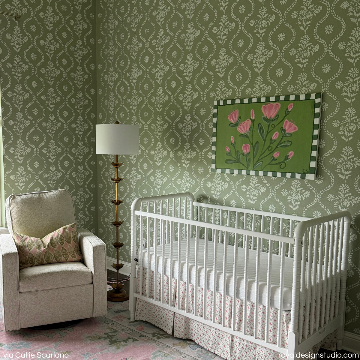 A styled nursery corner featuring a white wooden crib with a pink floral-patterned skirt, set against sage green floral stripe wall stencil pattern in a cottage style