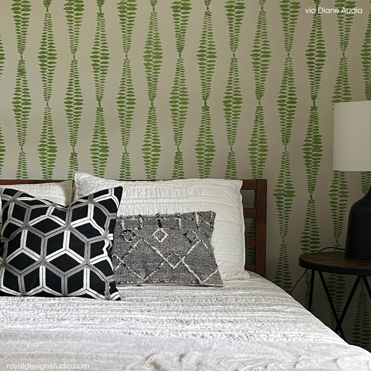 A styled bedroom scene featuring a bed with textured white bedding, two decorative throw pillows, and a wall covered in green and beige vertical leaf-patterned wall stencil design.