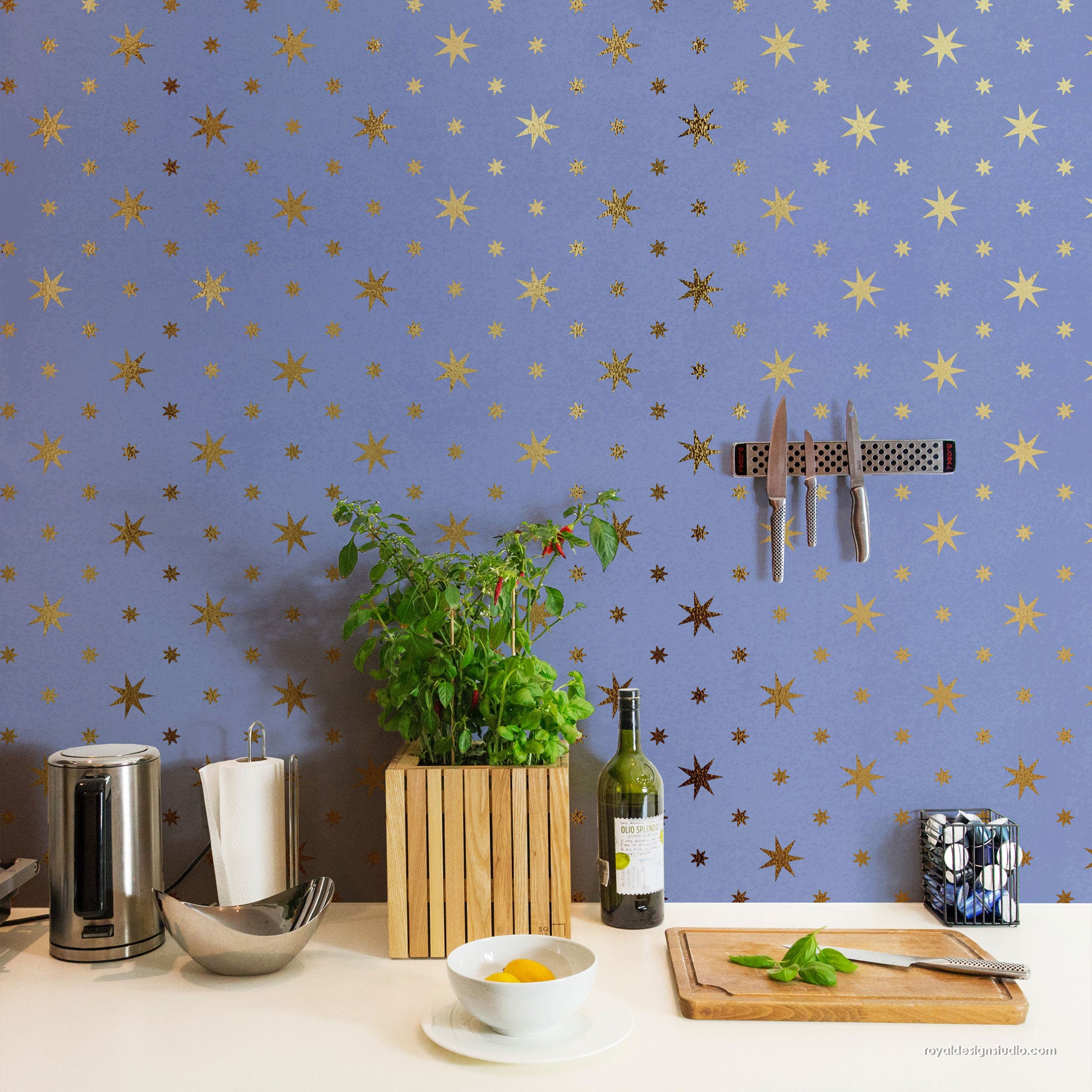 A styled kitchen countertop with a blue wall background patterned with gold stars. The scene includes a wooden slat planter with green herbs, a bottle of wine, a cutting board with fresh basil, a bowl of yellow food, and kitchen utensils, including a magnetic knife rack mounted on the wall.