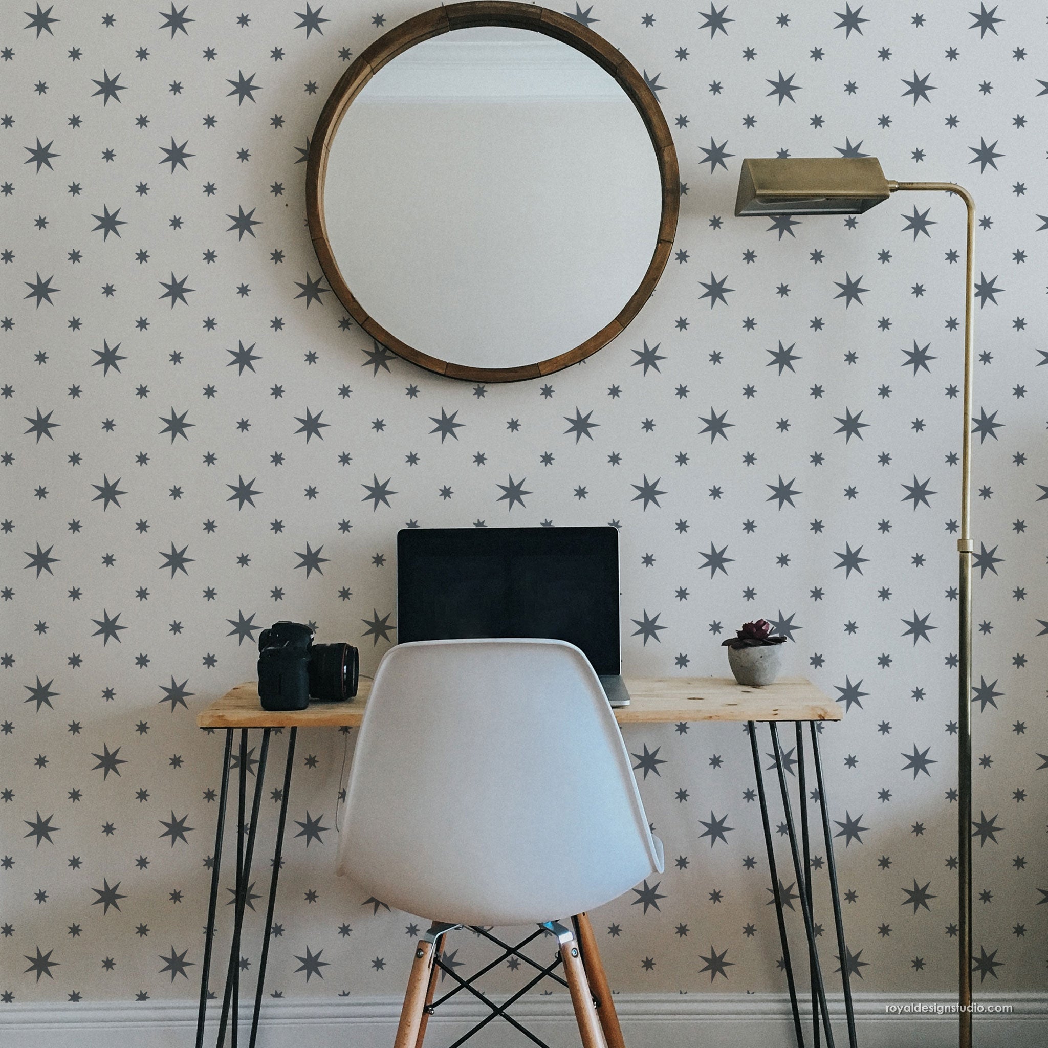 A styled home office corner featuring a light wood desk with black hairpin legs, a white molded chair, a laptop, a brass floor lamp, and a round wooden-framed mirror mounted on a wall with repeating gray starburst design.