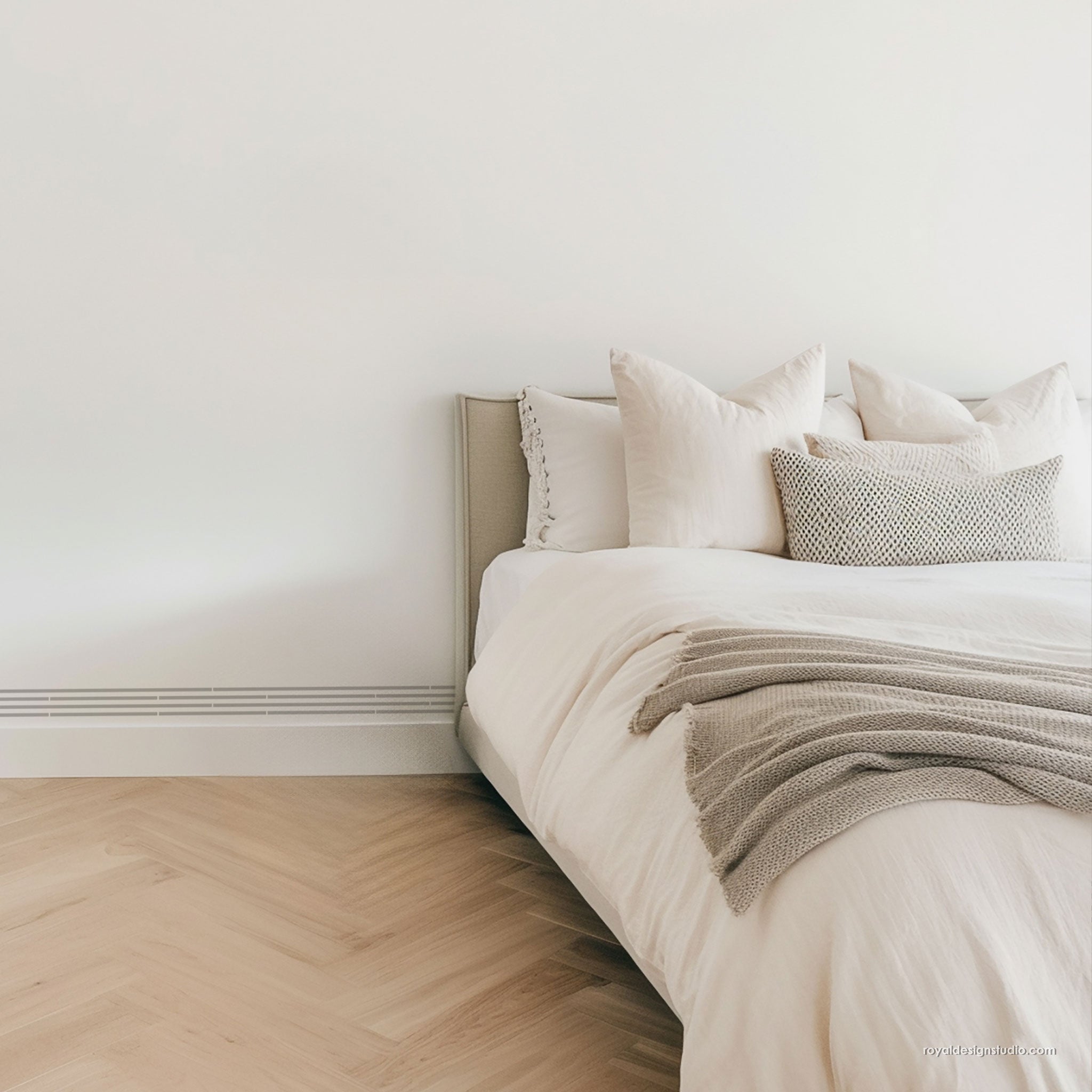 A minimalist bedroom corner showing a neatly made bed with white bedding, cream pillows, a textured gray throw blanket, a light wood headboard, pale wood herringbone flooring, and a white wall.