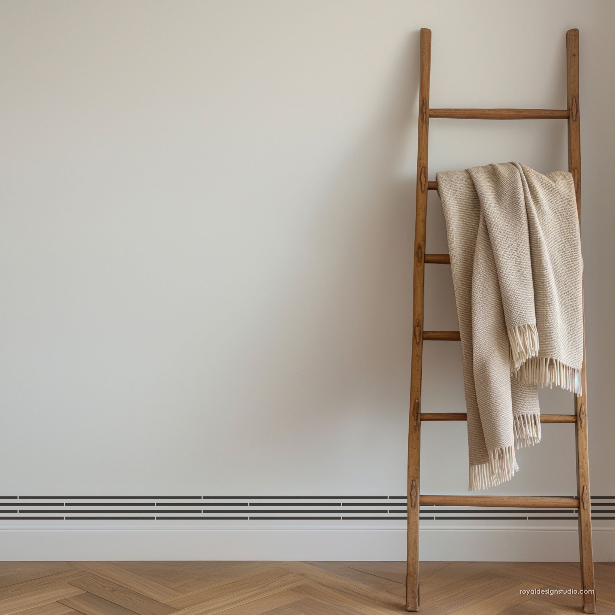 A rustic wooden ladder leans against a plain off-white wall on a light wood herringbone floor, with a cream-colored textured throw blanket draped over the middle rungs.