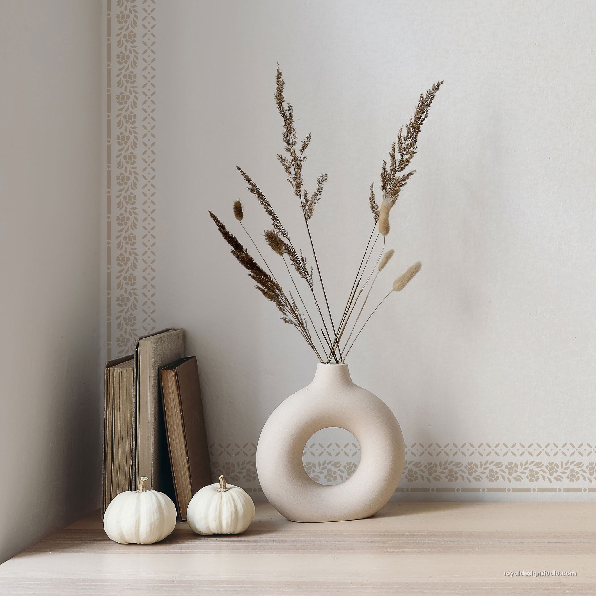 A matte ceramic donut-shaped vase in a warm cream color, filled with dried grasses, styled on a light wood surface next to two small white pumpkins and a stack of books against a neutral wall.