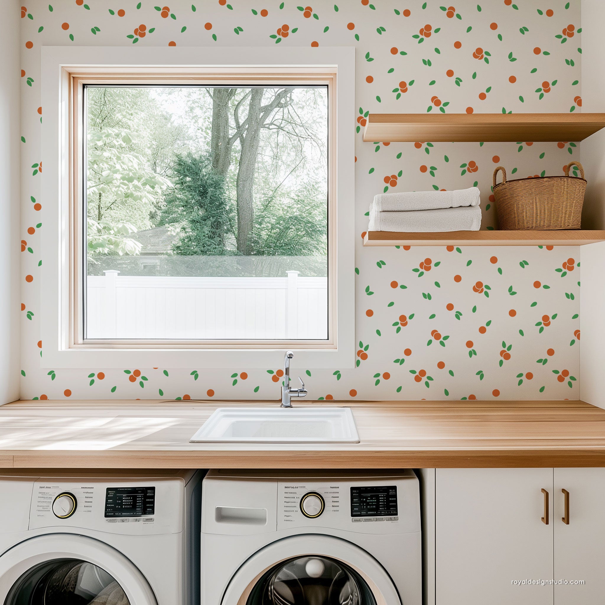 A bright, modern laundry room featuring a light wood countertop with a white sink, a window looking out to trees, and a white wall covered in an orange and green citrus pattern. Open wooden shelves on the right hold folded white towels and a wicker basket, while white laundry cabinets and washing machines are visible below.