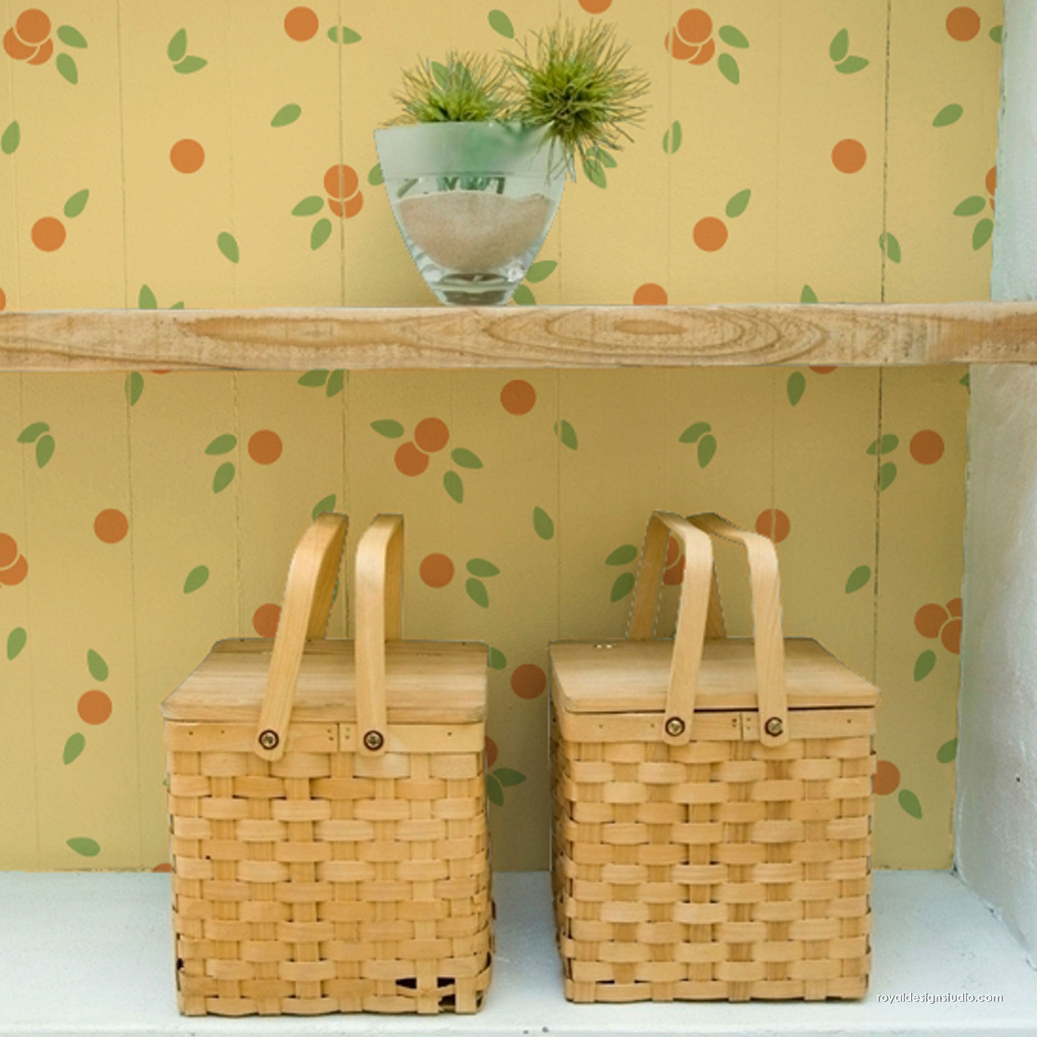Two woven wooden picnic baskets with light wood handles sit on a white shelf against a yellow wall with an orange and green fruit pattern. A small glass planter with green grass sits on a wooden shelf above.