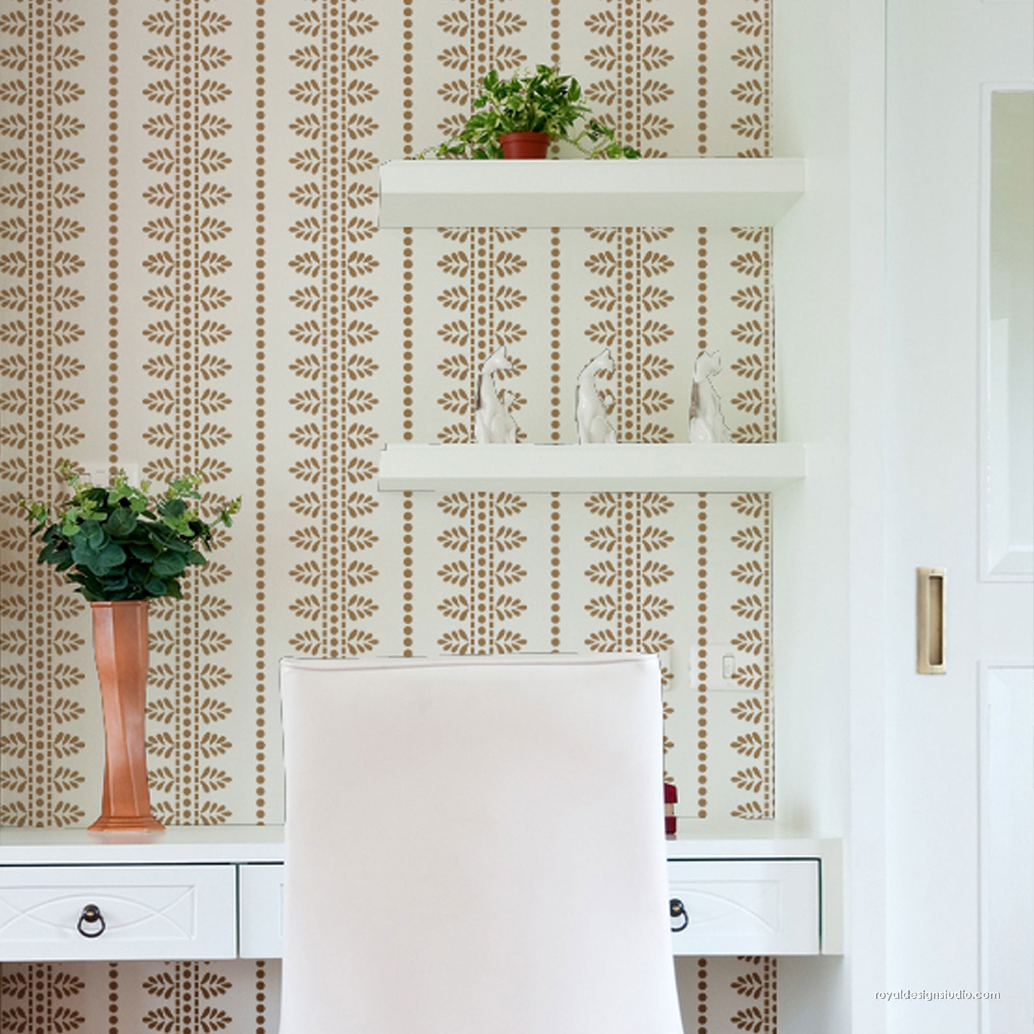 A styled interior scene showing a white desk with a white upholstered chair in front, white floating shelves, and patterned wall. A terracotta vase with greenery sits on the desk, and small decorative objects are arranged on the shelves.