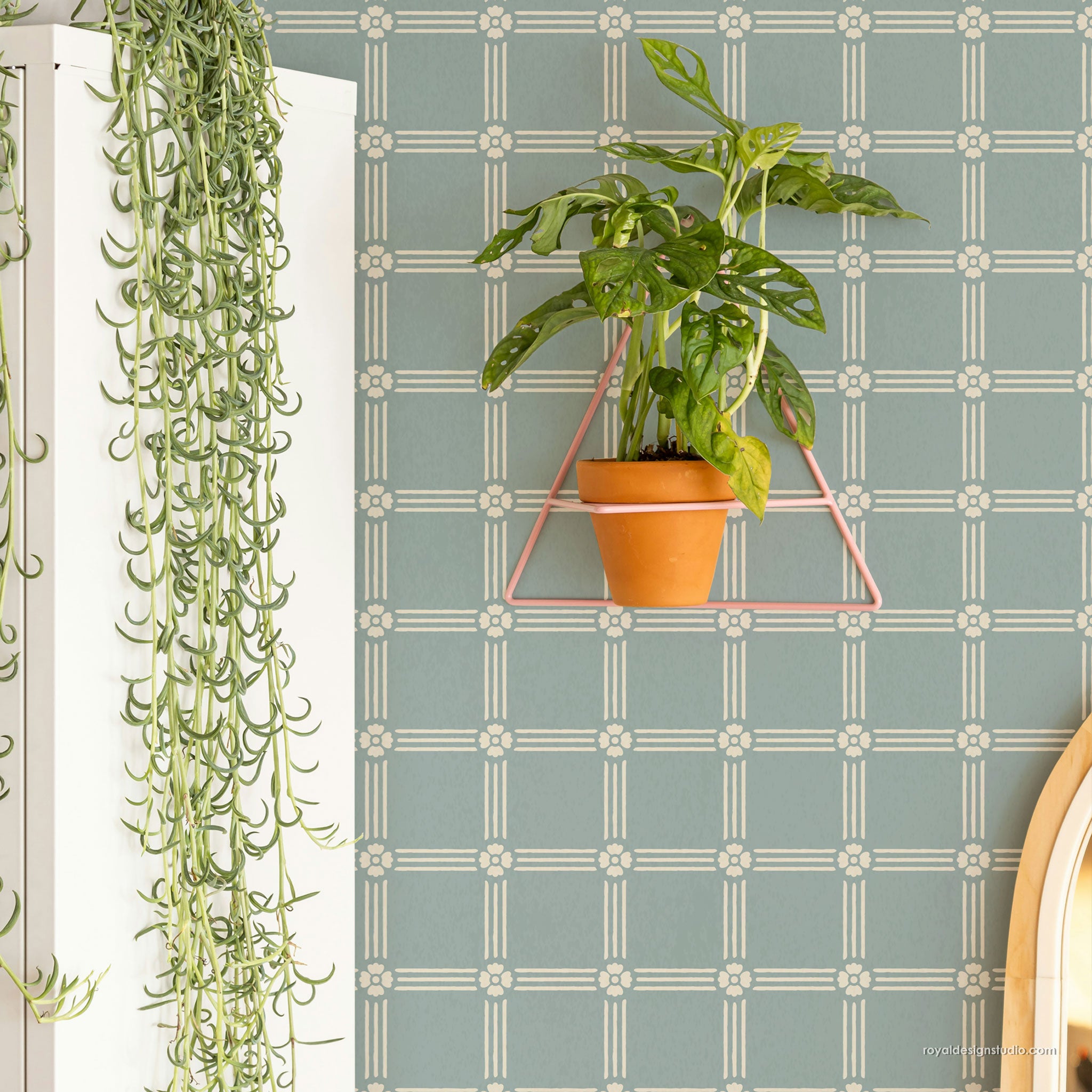 A potted green plant in a terracotta pot hanging from a pink triangular metal rack against a muted blue-green patterned wall. A white cabinet with a trailing green plant is visible on the left.