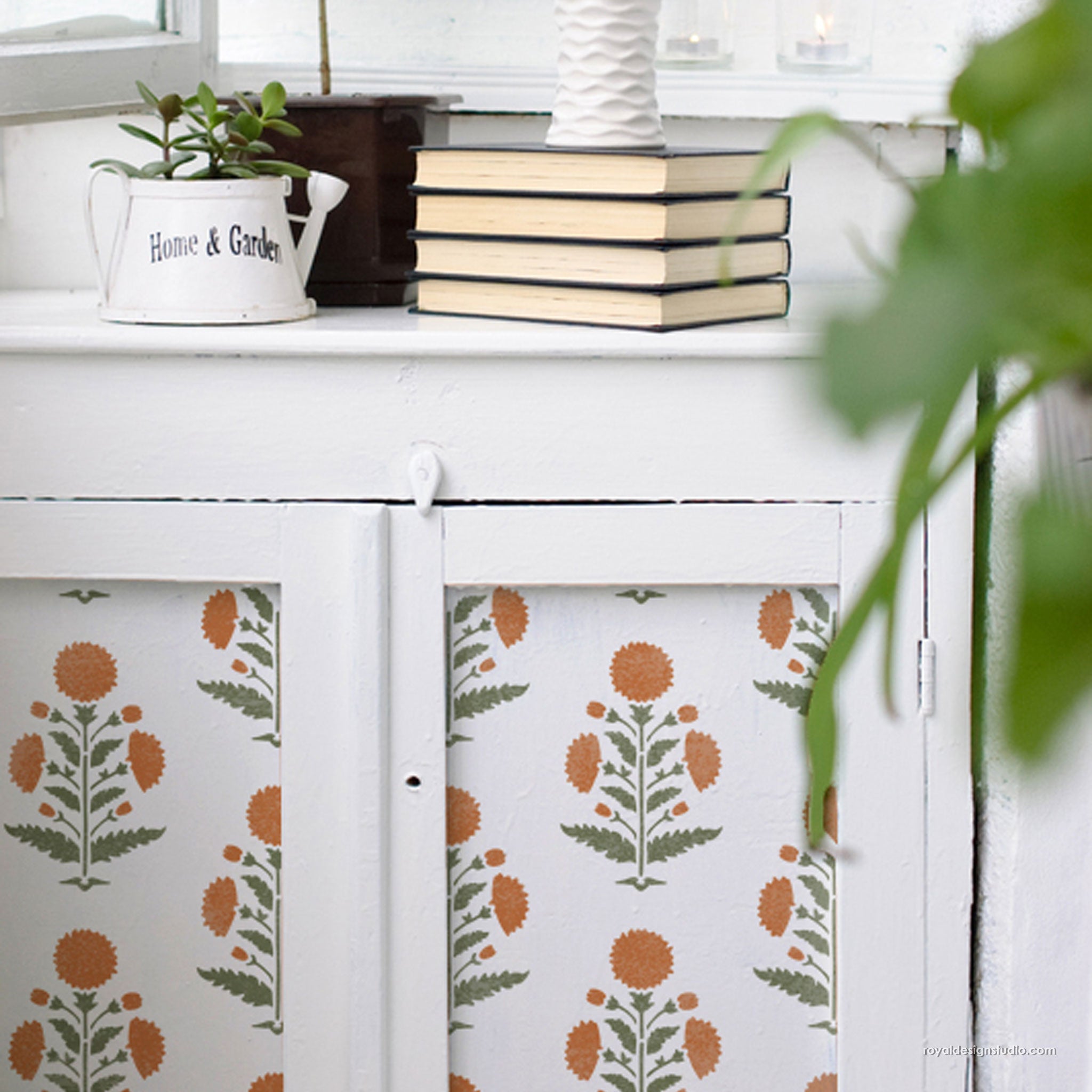 A white cabinet with floral-patterned doors in orange and green, styled in a bright room with books, a small potted plant, and a vase on top.
