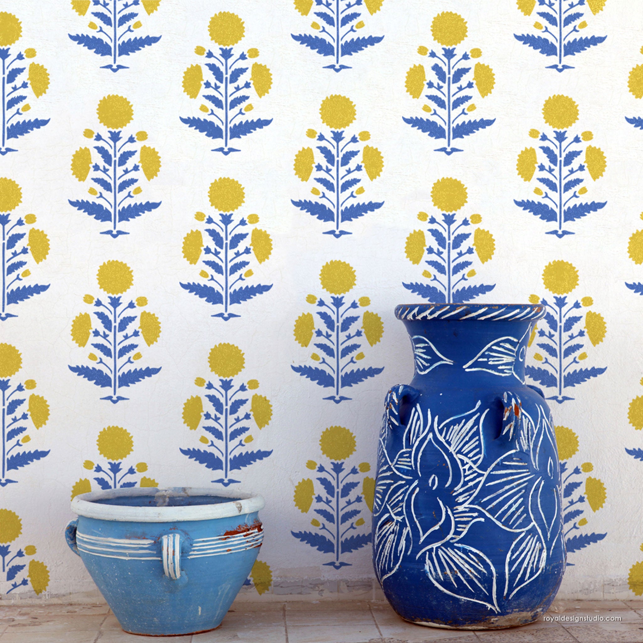 A blue ceramic vase with white floral line art stands next to a light blue ceramic pot against a white wall covered in a repeating blue and yellow floral block print pattern.