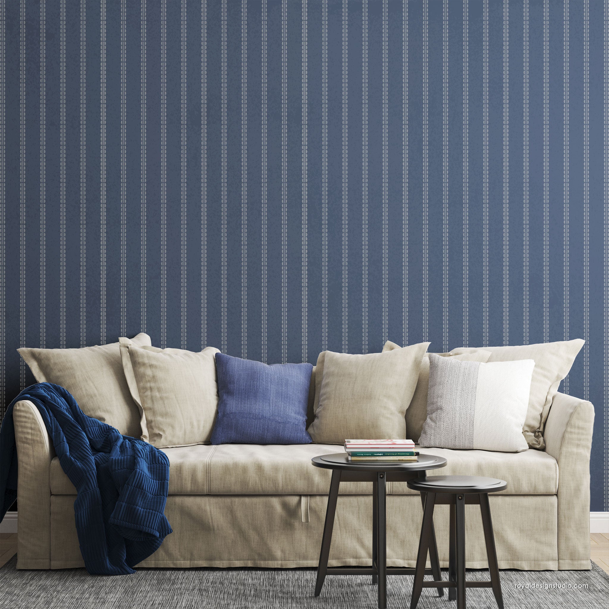A styled living room scene featuring a beige slipcovered sofa with multiple neutral and blue throw pillows, a navy blue throw blanket draped over the arm, and a small round side table in front. The wall behind is a vertical navy wall with a light blue geometric stripe pattern.