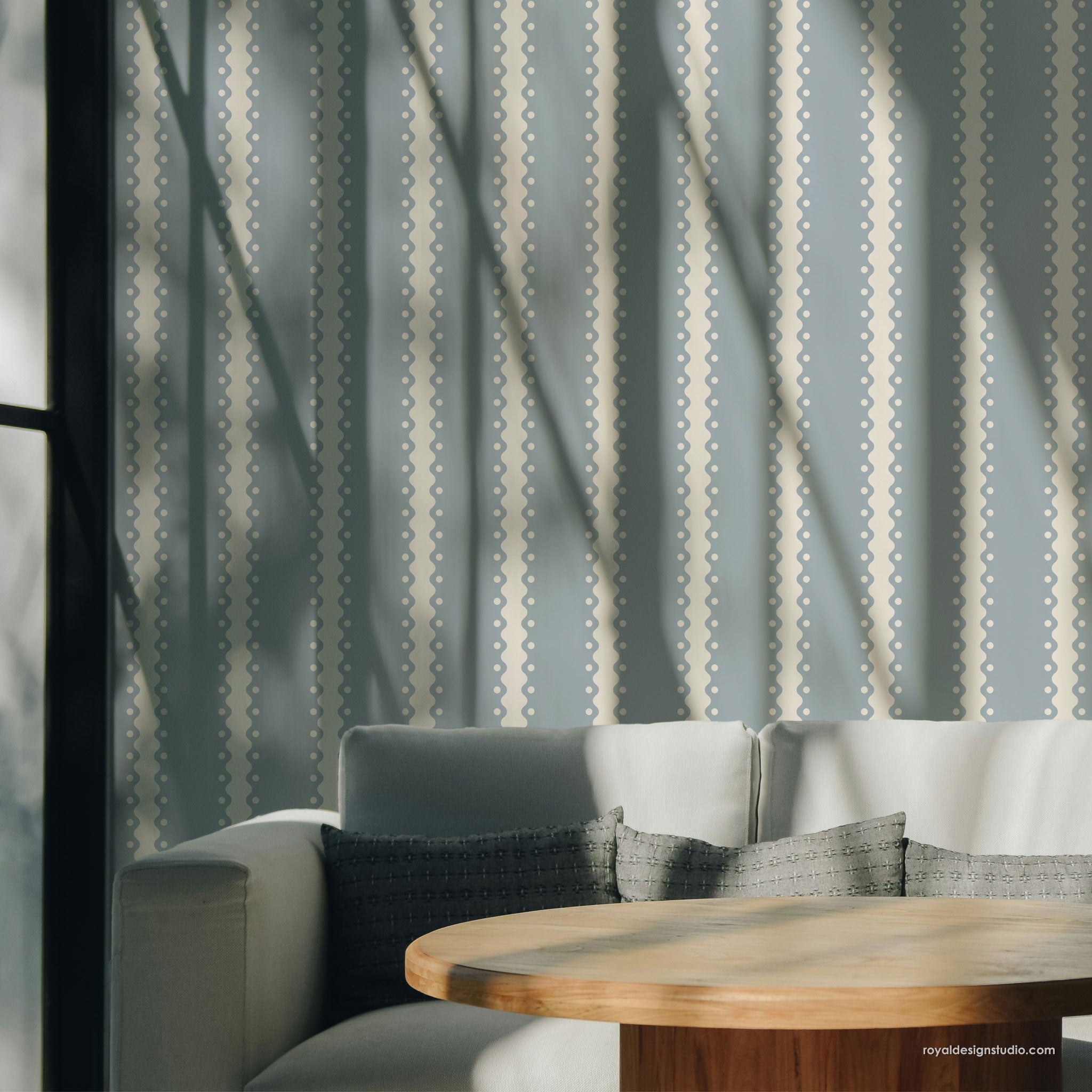 A styled interior scene showing a light gray sofa with patterned gray pillows and a round wooden coffee table, set against a wall with vertical blue-gray and cream patterned wall and strong diagonal sunlight shadows.